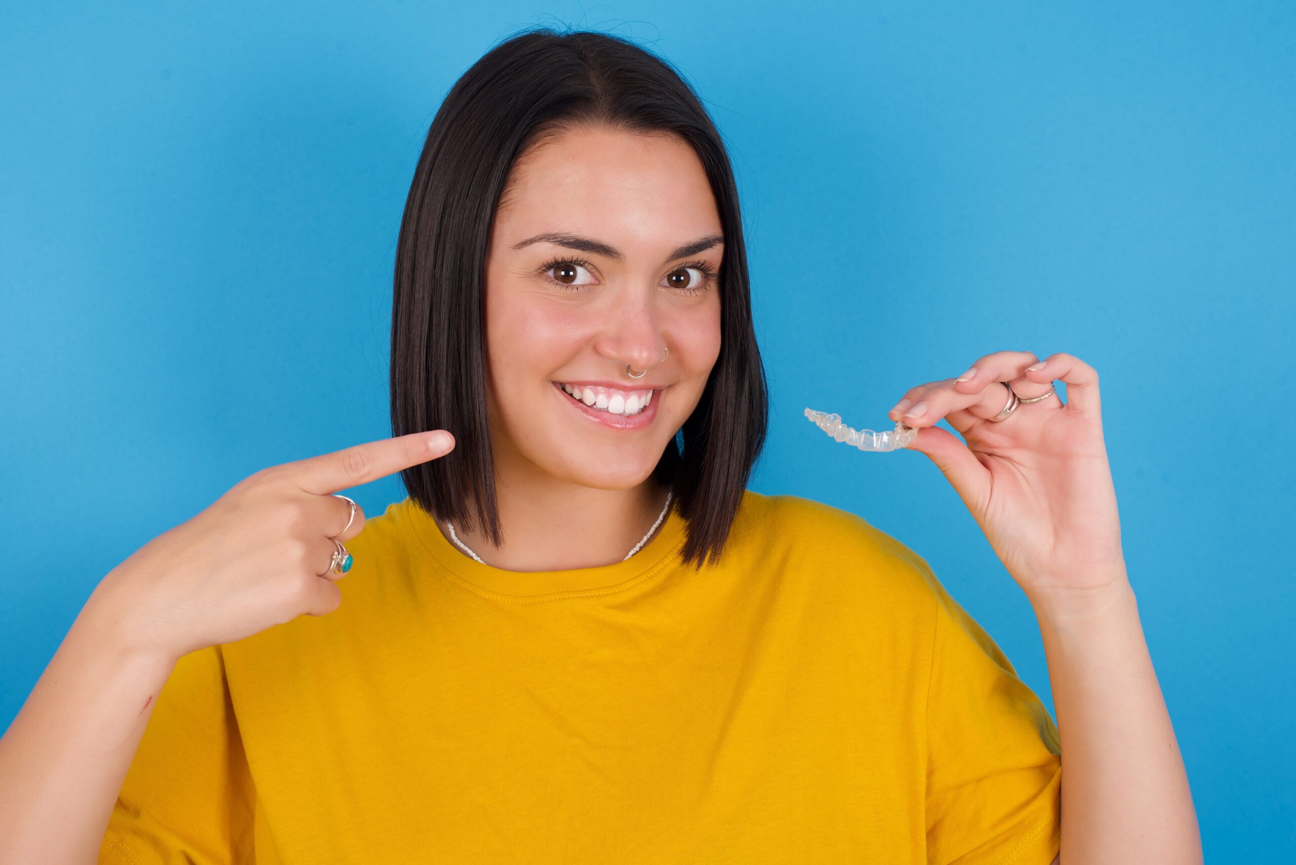 MODEL holding an invisible aligner and pointing to her perfect straight teeth. Dental healthcare and confidence concept.