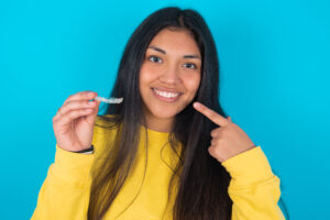MODEL holding an invisible aligner and pointing to her perfect straight teeth. Dental healthcare and confidence concept.